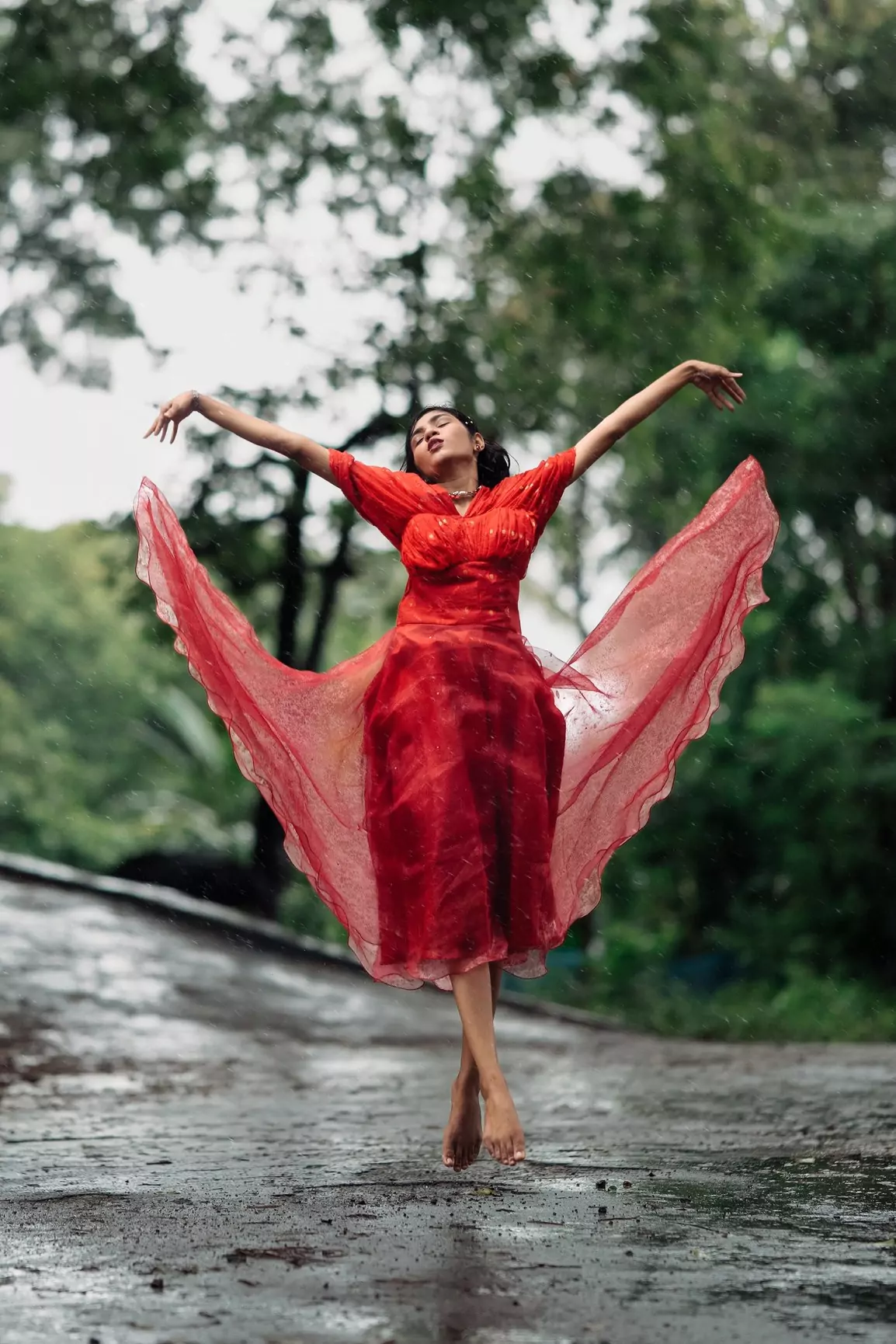 Striking pose with arms wide open, red dress fanned out on a rain-slicked road