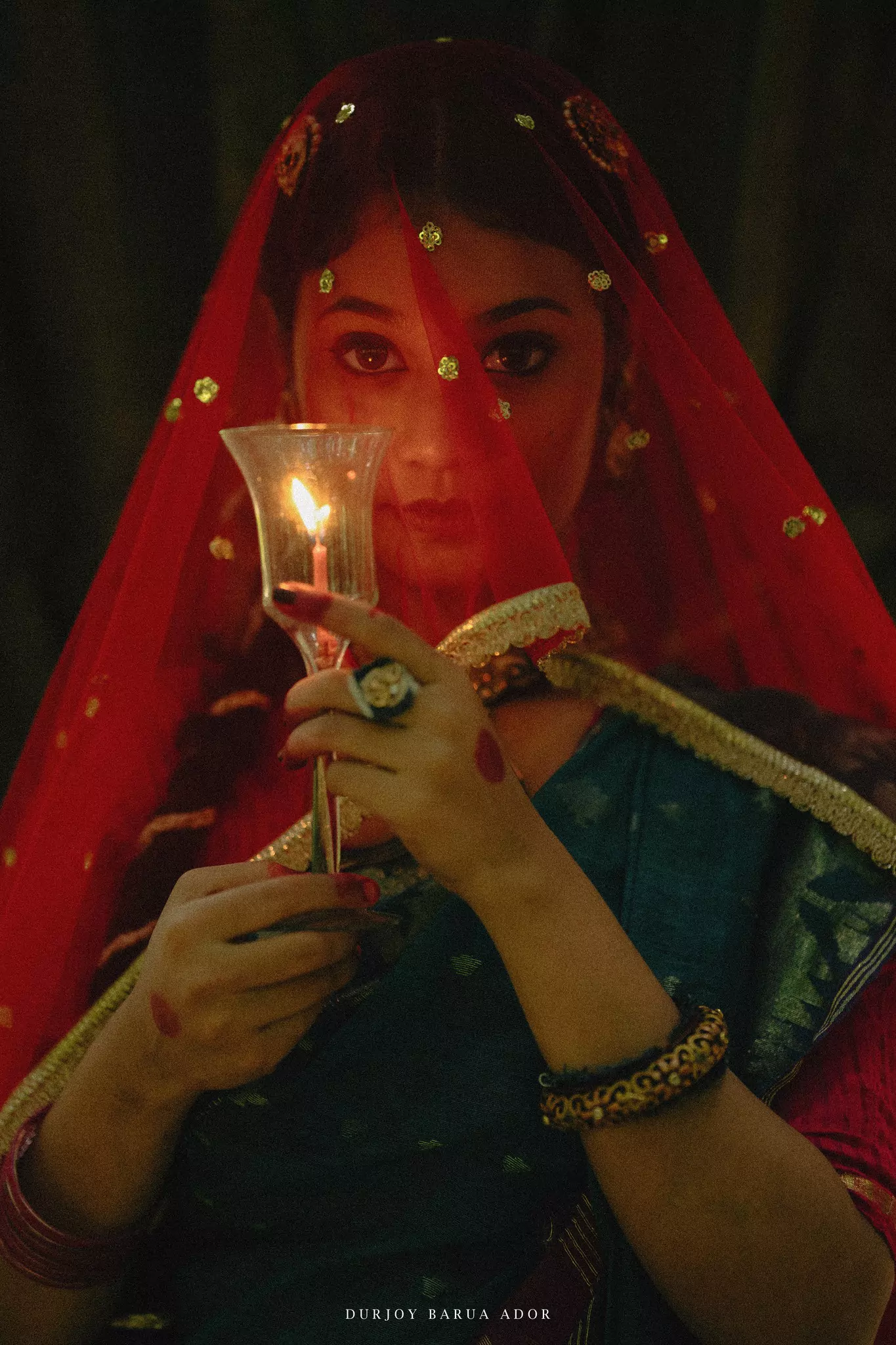 Close-up of woman in red veil and blue saree holding a diya with dramatic lighting.
